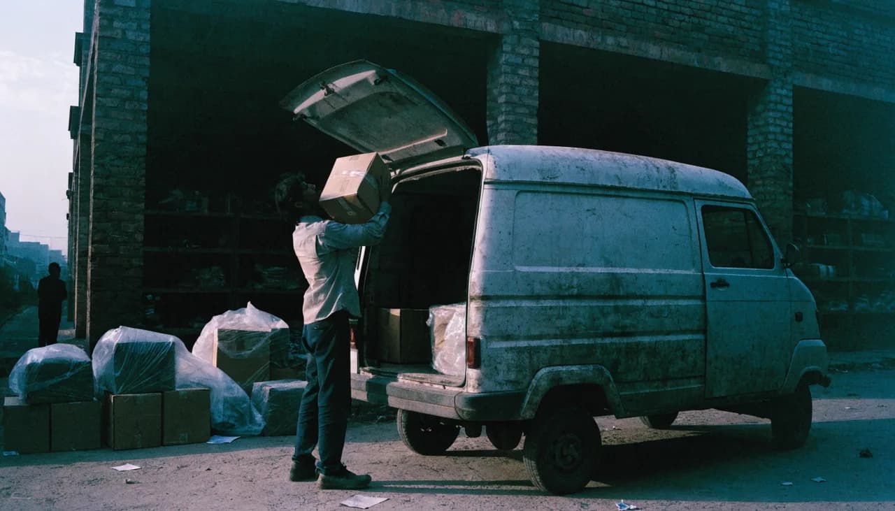 A driver lifting a sealed carton into the back of a delivery van outside a godown at first light, parcels stacked beside him.